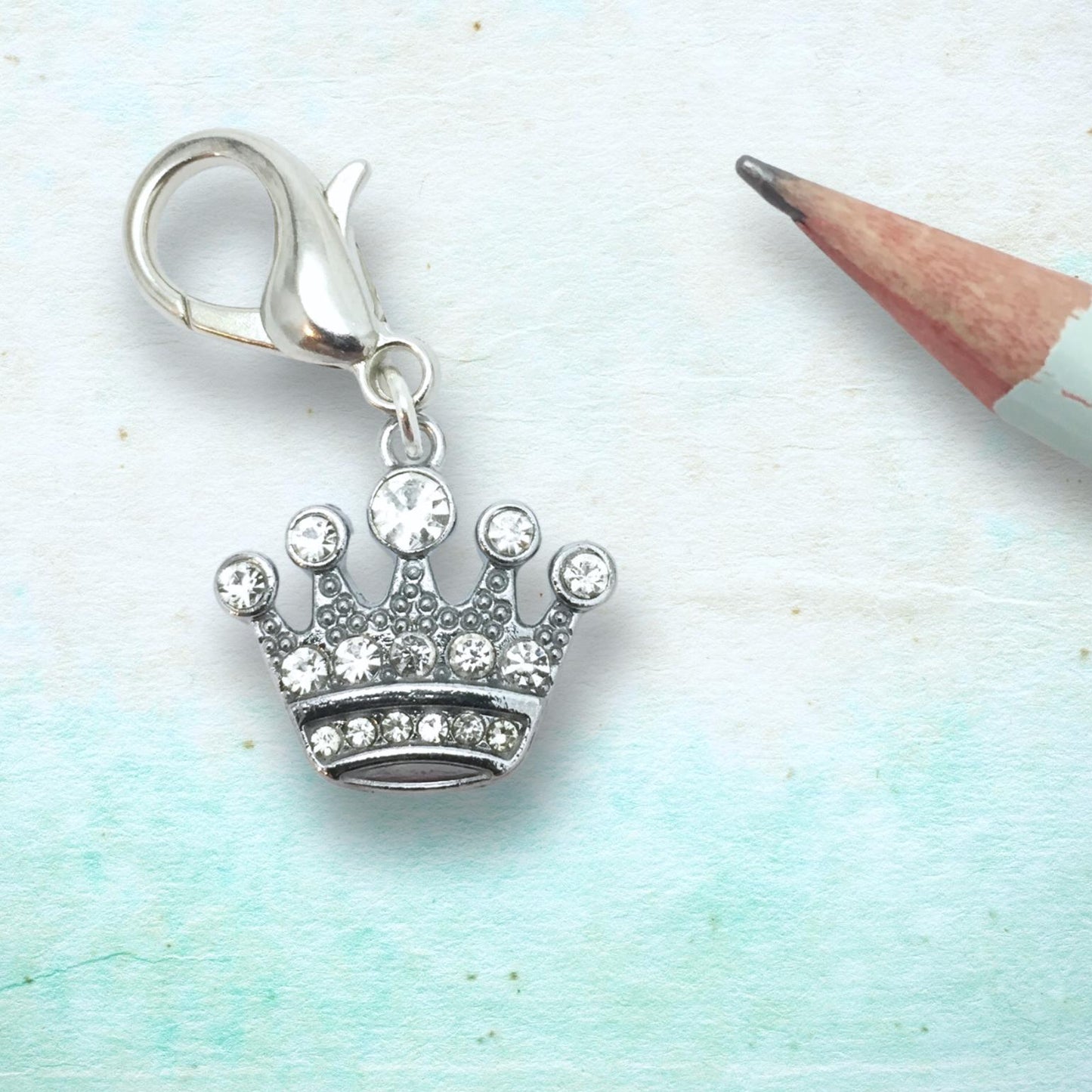 A silver crown charm with clear rhinestones and a lobster clasp, displayed on a light blue textured background next to a pencil.