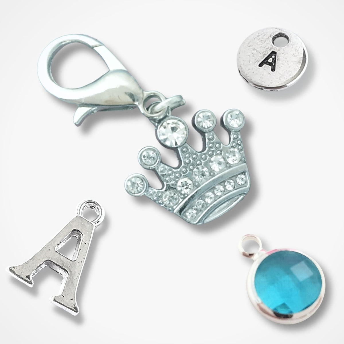 Silver jewelry charms on a light gray background, including a crown with clear rhinestones, a silver letter A charm, a round silver disc engraved with the letter A, and a round turquoise-blue faceted gemstone bezel.