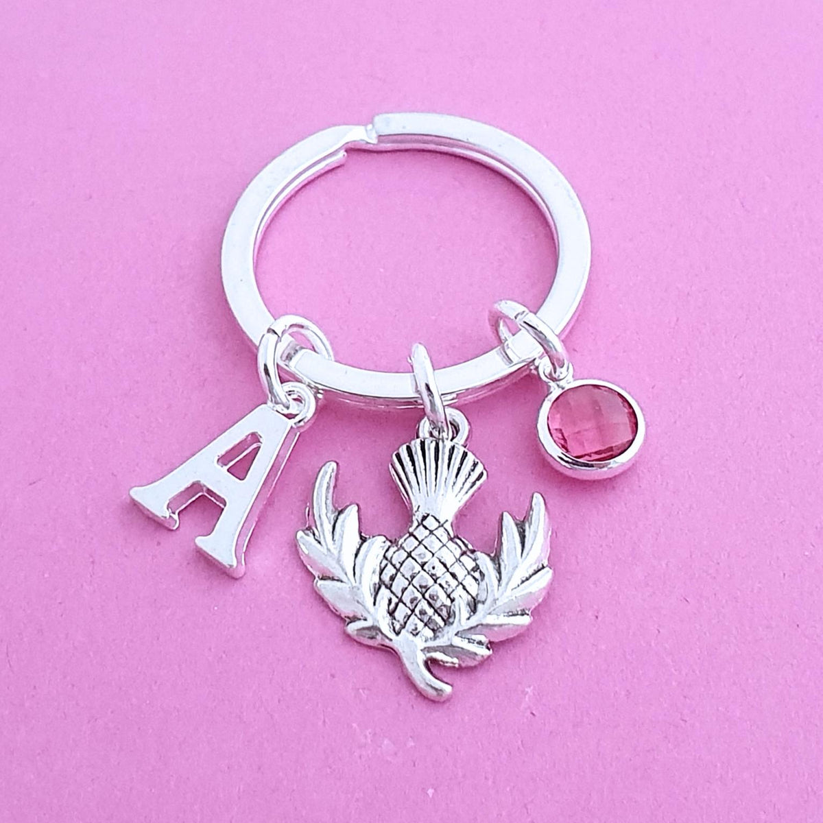 A silver keychain with a split ring, featuring a letter 'A' charm, a thistle emblem charm, and a small round pink gemstone charm, displayed on a solid pink background.