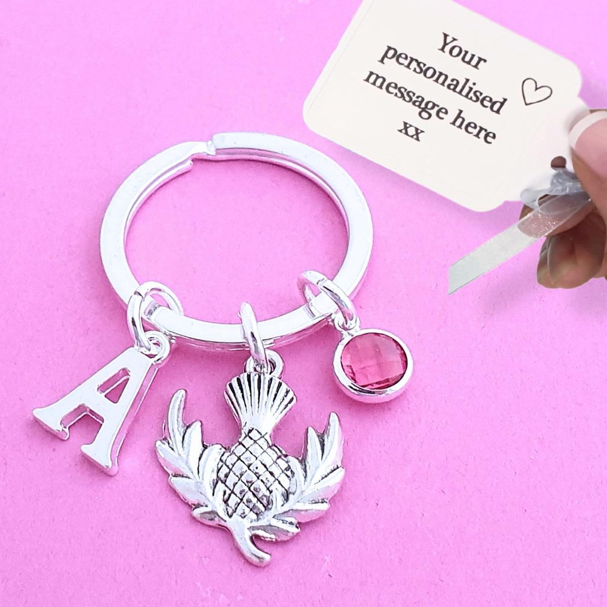 A silver key ring charm set featuring a letter 'A', a thistle, and a pink gemstone bead, shown on a pink background with a hand holding a customization card.