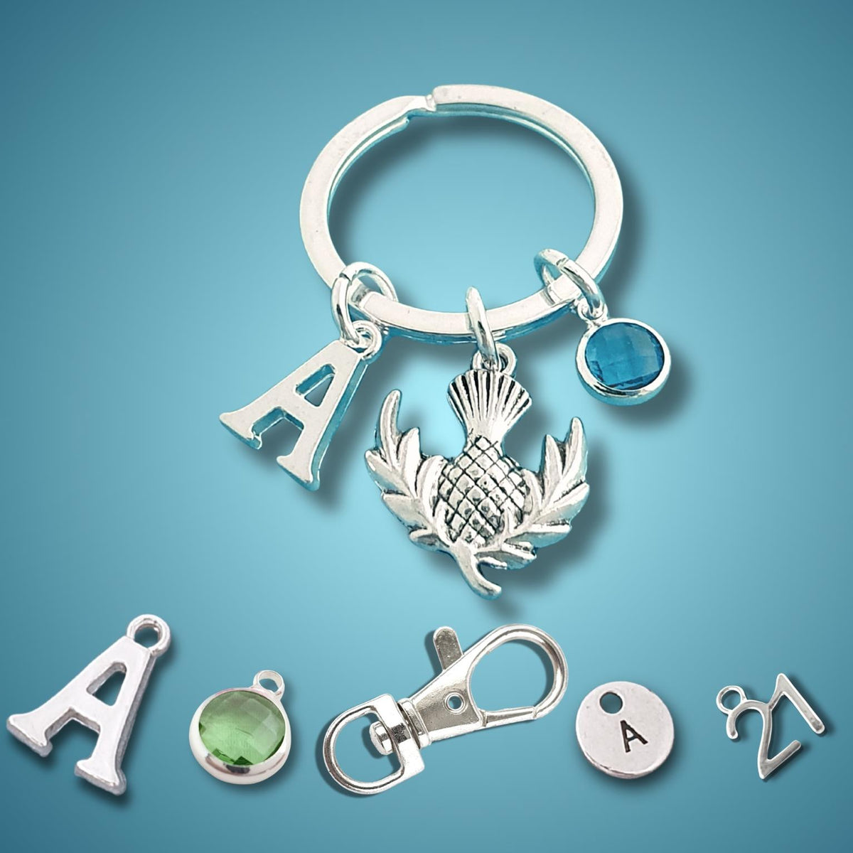 A silver keychain set featuring a split ring with charms including a silver thistle, a letter 'A', a blue stone, and several loose matching charms on a blue gradient background.