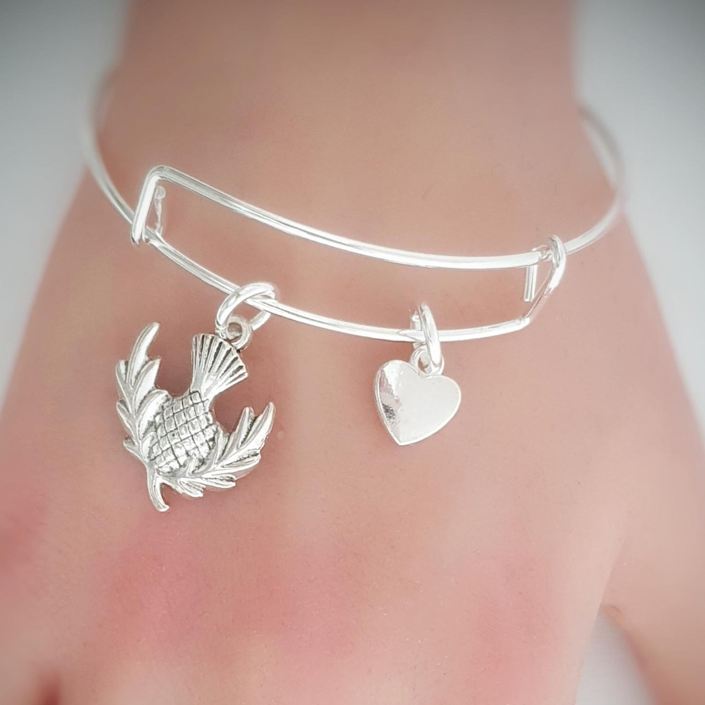 Silver bracelet with thistle and heart charms on a wrist against a neutral background