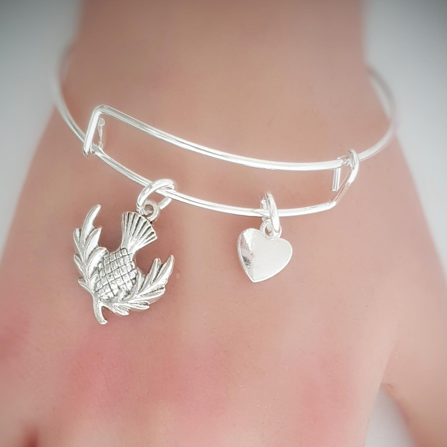 Silver bracelet with thistle and heart charms on a wrist against a neutral background