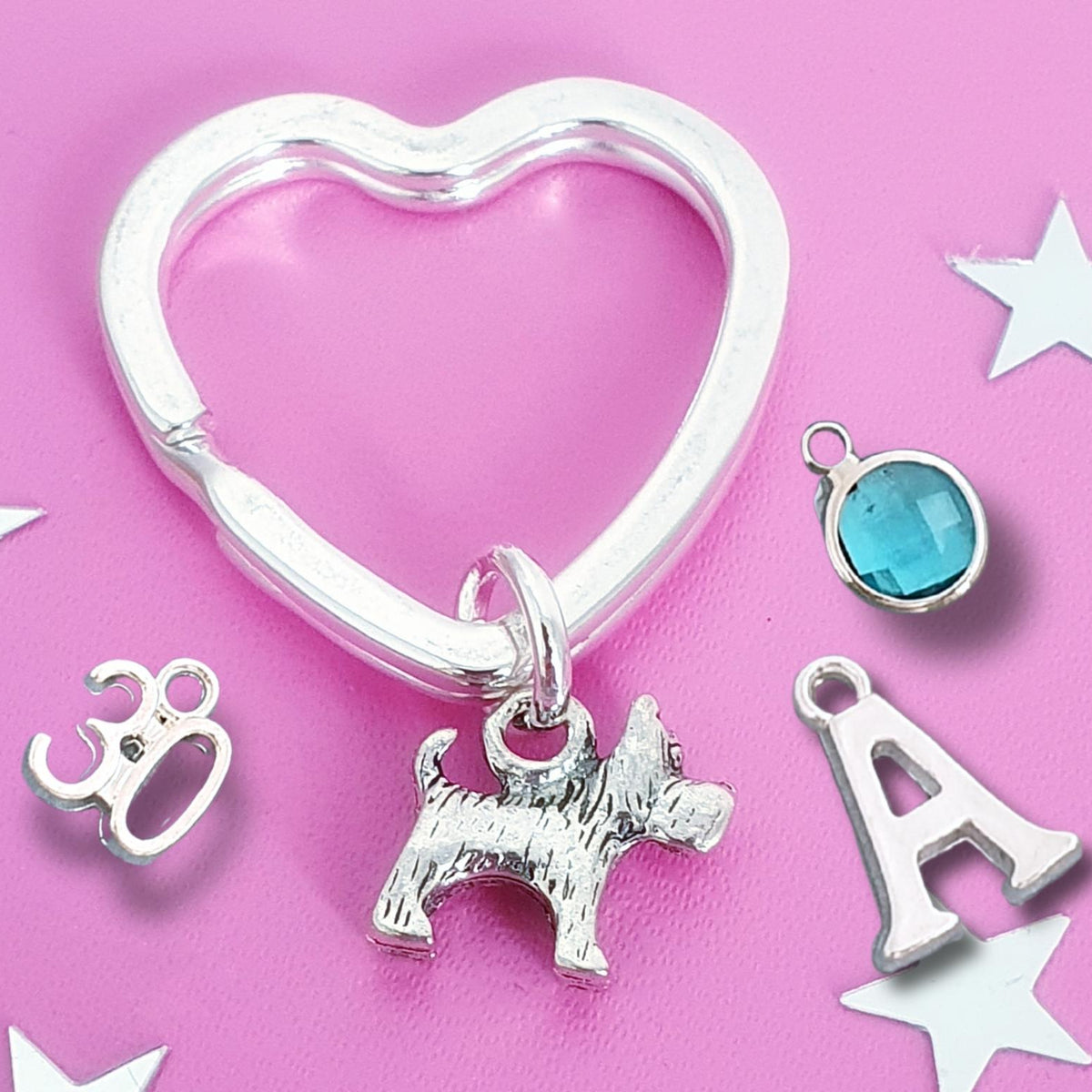 A silver heart-shaped key ring charm set displayed on a pink background with silver star cutouts. The set includes a textured Scottish Terrier dog charm, a letter 'A' initial charm, a '30' charm, and a round blue faceted gemstone charm.
