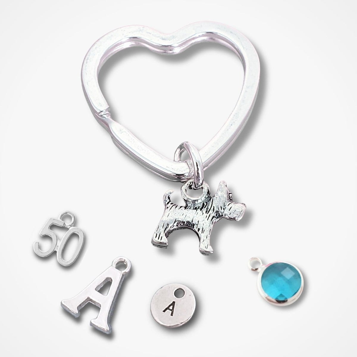 A silver heart-shaped key ring with dangling charms, including a small scotty dog silhouette, a letter A, a round engraved disc with an A, a blue faceted gemstone, and a silver '50' charm, displayed on a light gray background.