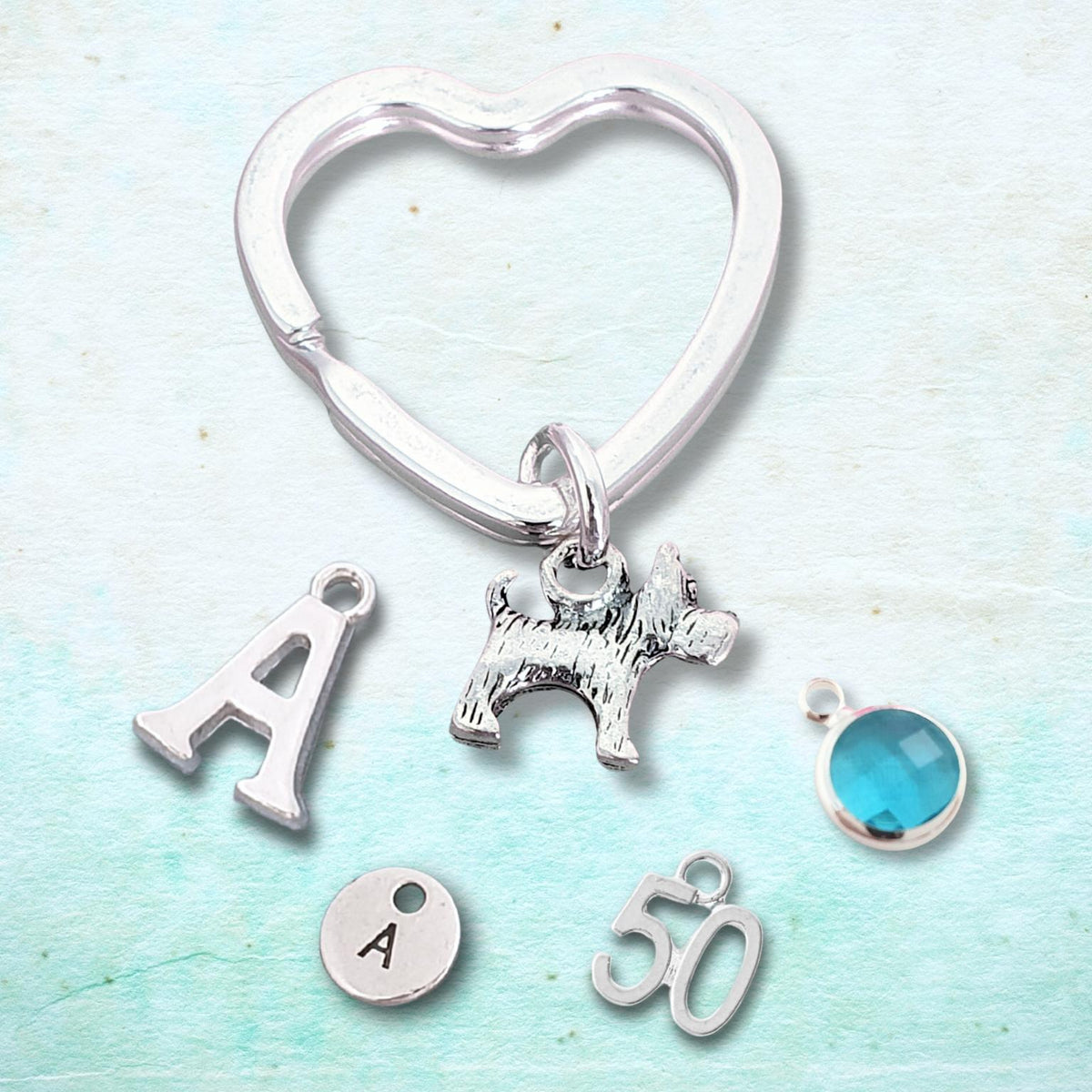 A silver heart-shaped key ring charm set with multiple personalized charms, including a scottie dog silhouette, a letter A, a round engraved disc with the letter A, a number 50, and a round turquoise-blue stone charm, displayed on a textured light blue background.