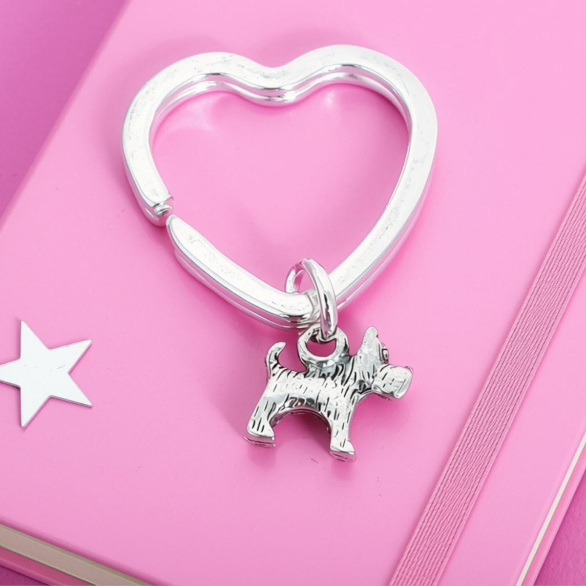 A silver heart-shaped key ring with a small scotty dog charm, displayed on a pink notebook.