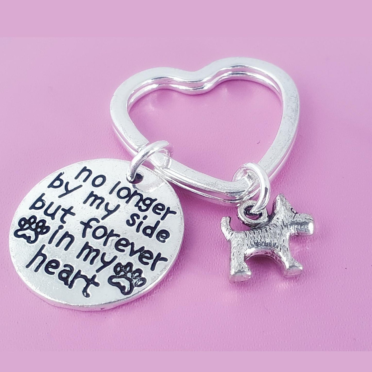 A silver-toned metal keychain featuring a heart-shaped ring, a round engraved disc with the message 'no longer by my side but forever in my heart' and paw prints, and a small dog charm, displayed on a solid pink background.