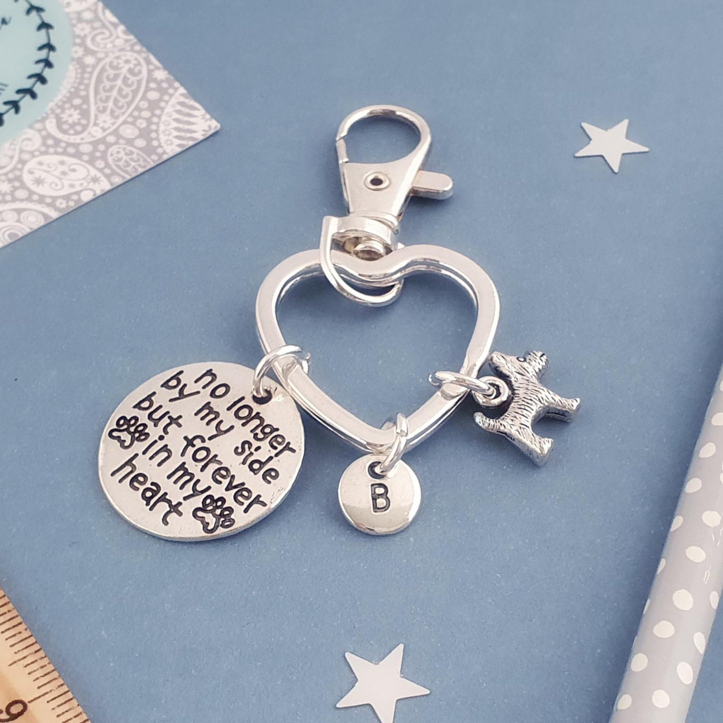 A silver heart-shaped keychain with charms, including a round engraved disc that reads 'no longer by my side but forever in my heart', a small letter B disc charm, and a dog silhouette charm, displayed on a blue background with decorative props.