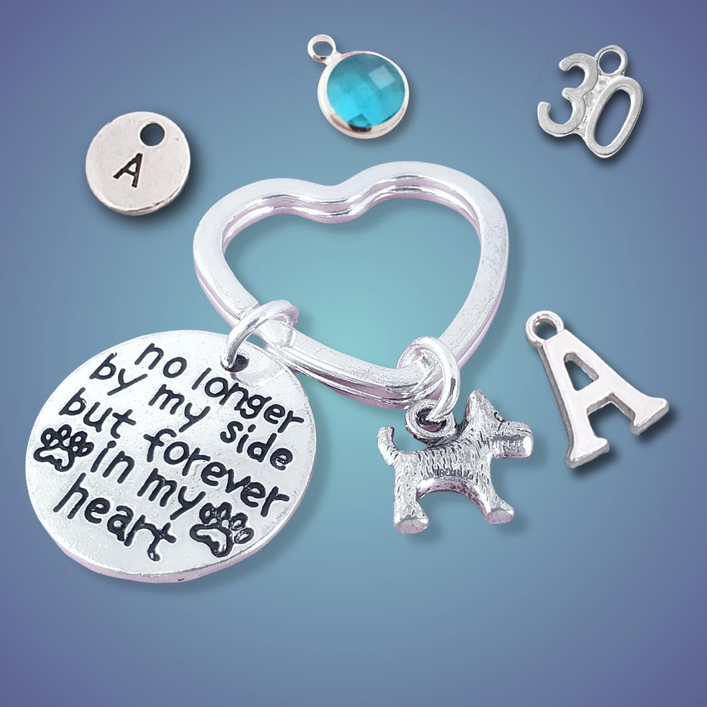 A silver memorial keychain set featuring a heart-shaped ring, a round engraved disc with the text 'no longer by my side but forever in my heart', a small dog charm, an 'A' initial charm, a '30' charm, and a round bezel-set blue gemstone charm, all arranged on a blue gradient background.