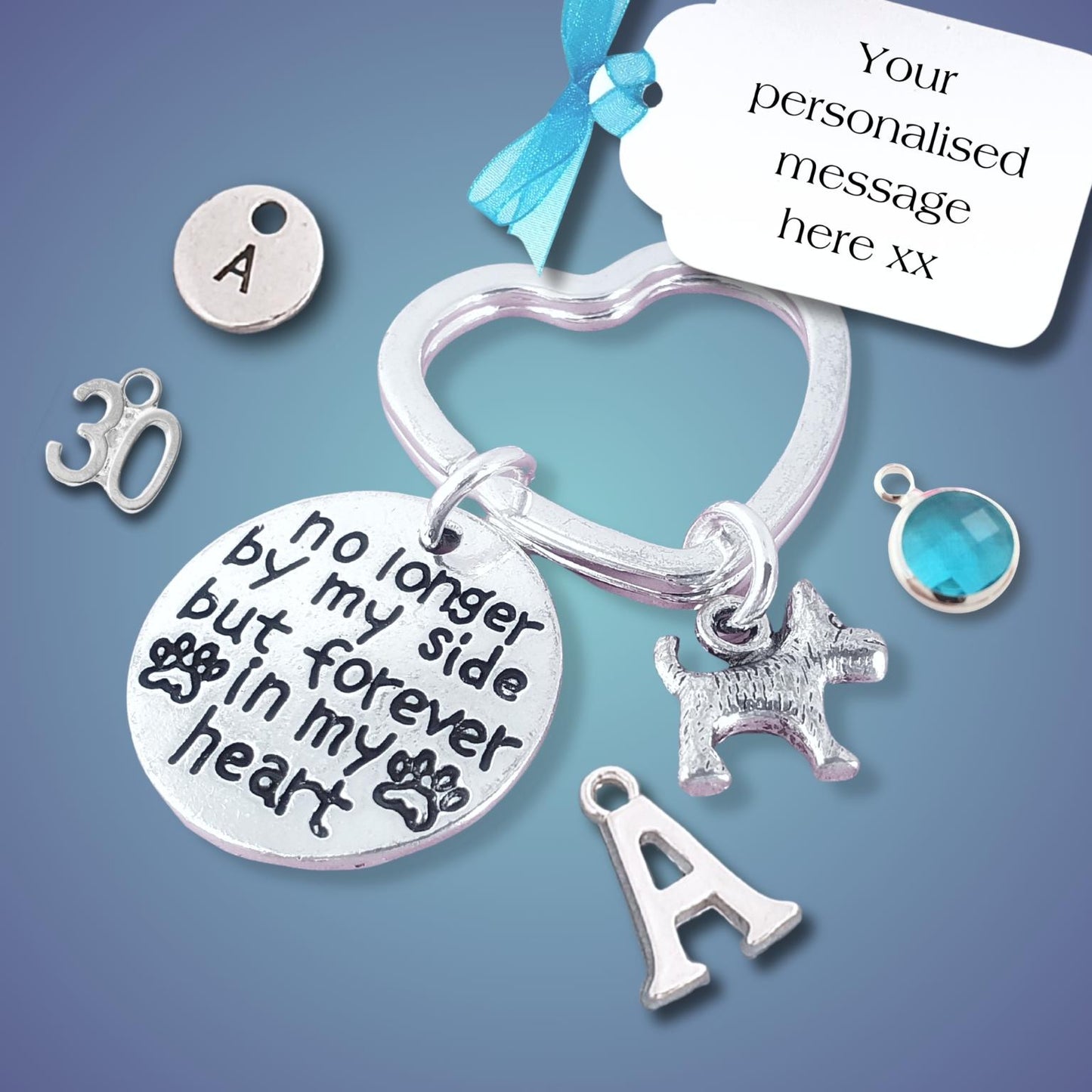 A silver memorial keychain set with charms, including a heart-shaped ring, a round disc engraved with 'no longer by my side but forever in my heart', a dog silhouette charm, an initial 'A' charm, a '30' charm, and a blue gemstone accent, shown with a white message tag on a blue gradient background.