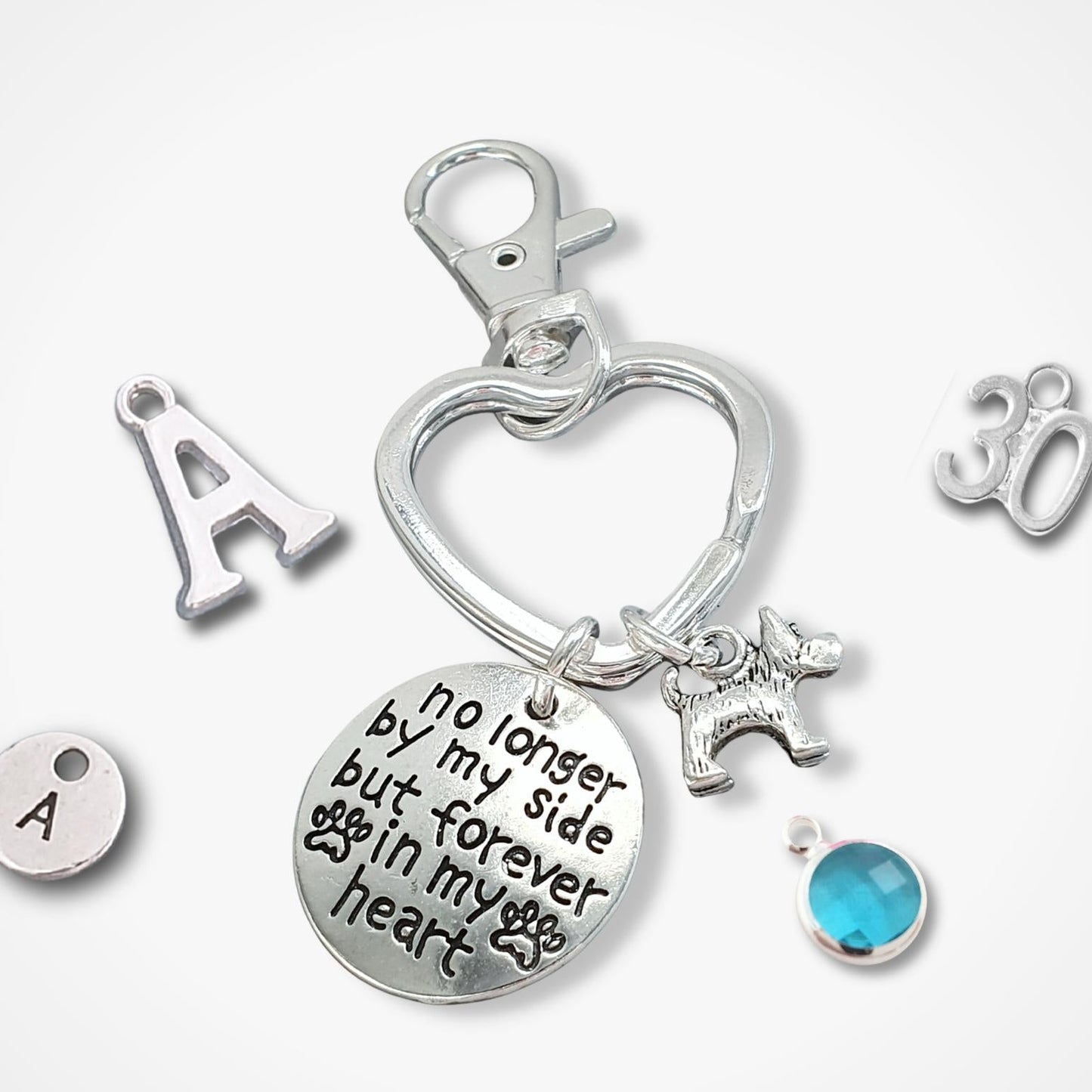 Silver memorial keychain charm set featuring a heart-shaped clasp, a round engraved disc with 'no longer by my side but forever in my heart', a dog silhouette charm, a blue faceted gemstone, and individual letter 'A' and number '30' charms on a white background.