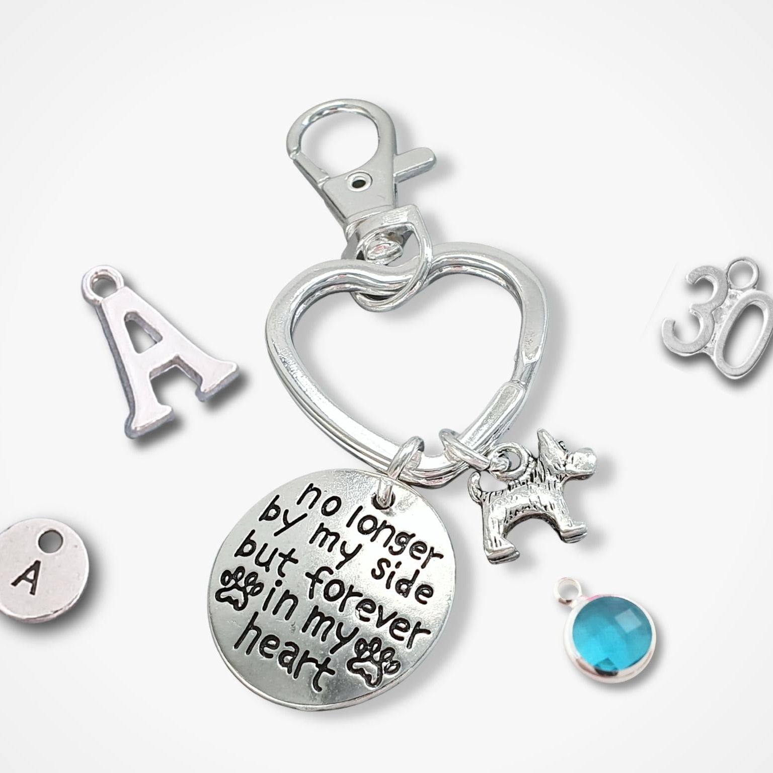 Silver memorial keychain charm set featuring a heart-shaped clasp, a round engraved disc with 'no longer by my side but forever in my heart', a dog silhouette charm, a blue faceted gemstone, and individual letter 'A' and number '30' charms on a white background.