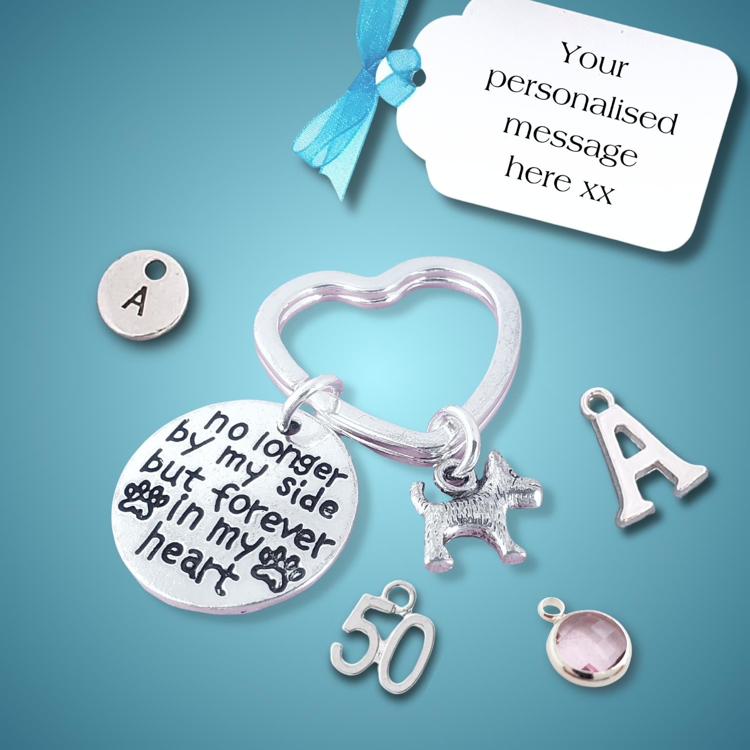Silver personalized pet memorial keychain with heart-shaped ring, engraved round disc reading 'no longer by my side but forever in my heart', dog charm, letter 'A' charms, number '50' charm, and a pink stone bezel, shown with a blue ribbon and white tag on a blue gradient background.