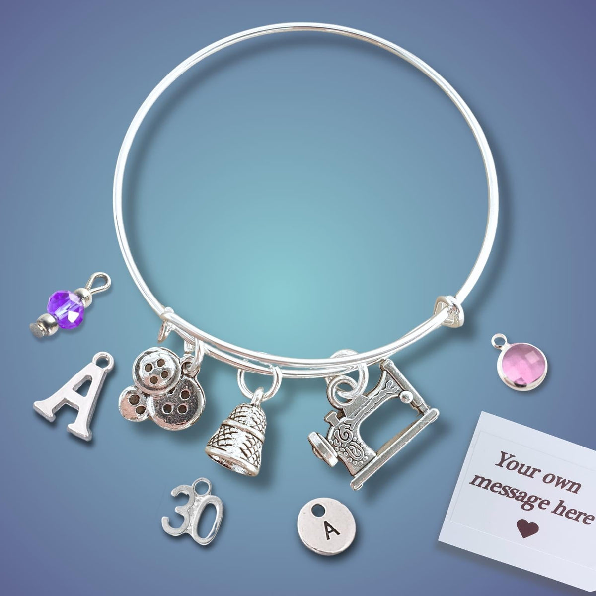 A silver bangle bracelet with multiple charms, including letters, a sewing machine, a thimble, and a button, displayed on a blue gradient background with a customization tag.