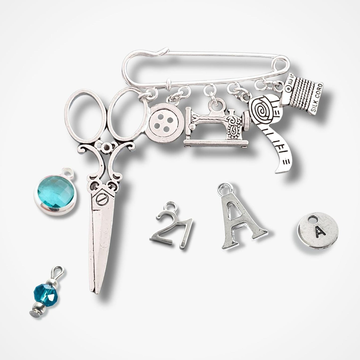 Brooch with sewing themed silver charms including scissors, sewing machine, buttons, and letters on a white background