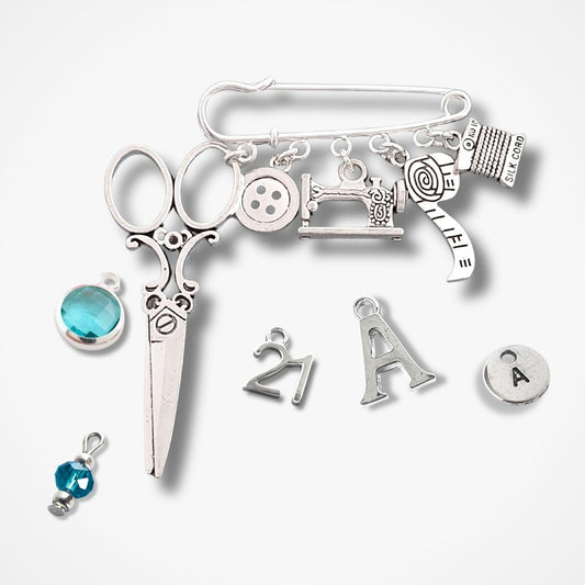 Brooch with sewing themed silver charms including scissors, sewing machine, buttons, and letters on a white background