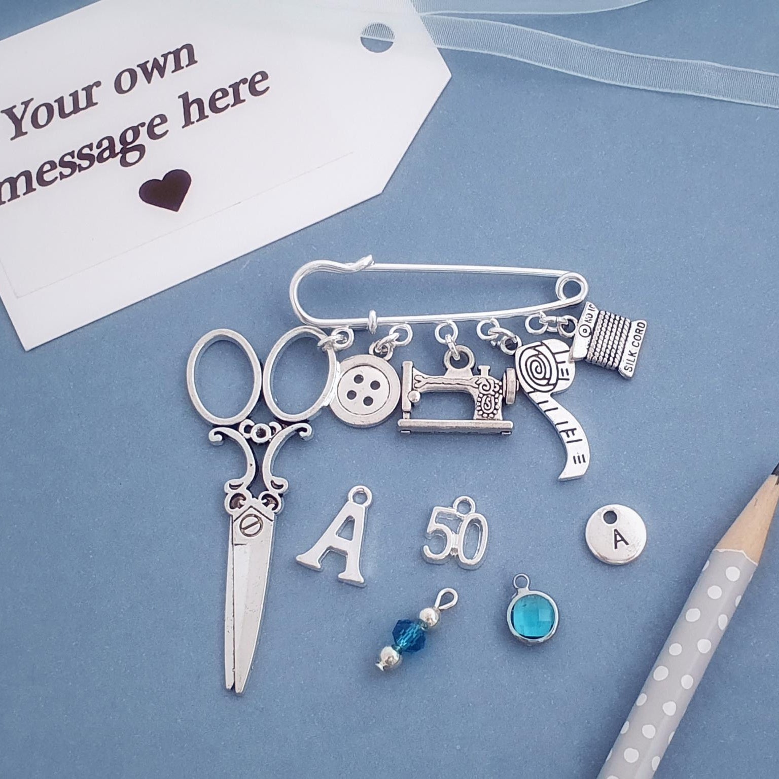 Collection of silver sewing-themed charms on a blue background with a pencil and card.