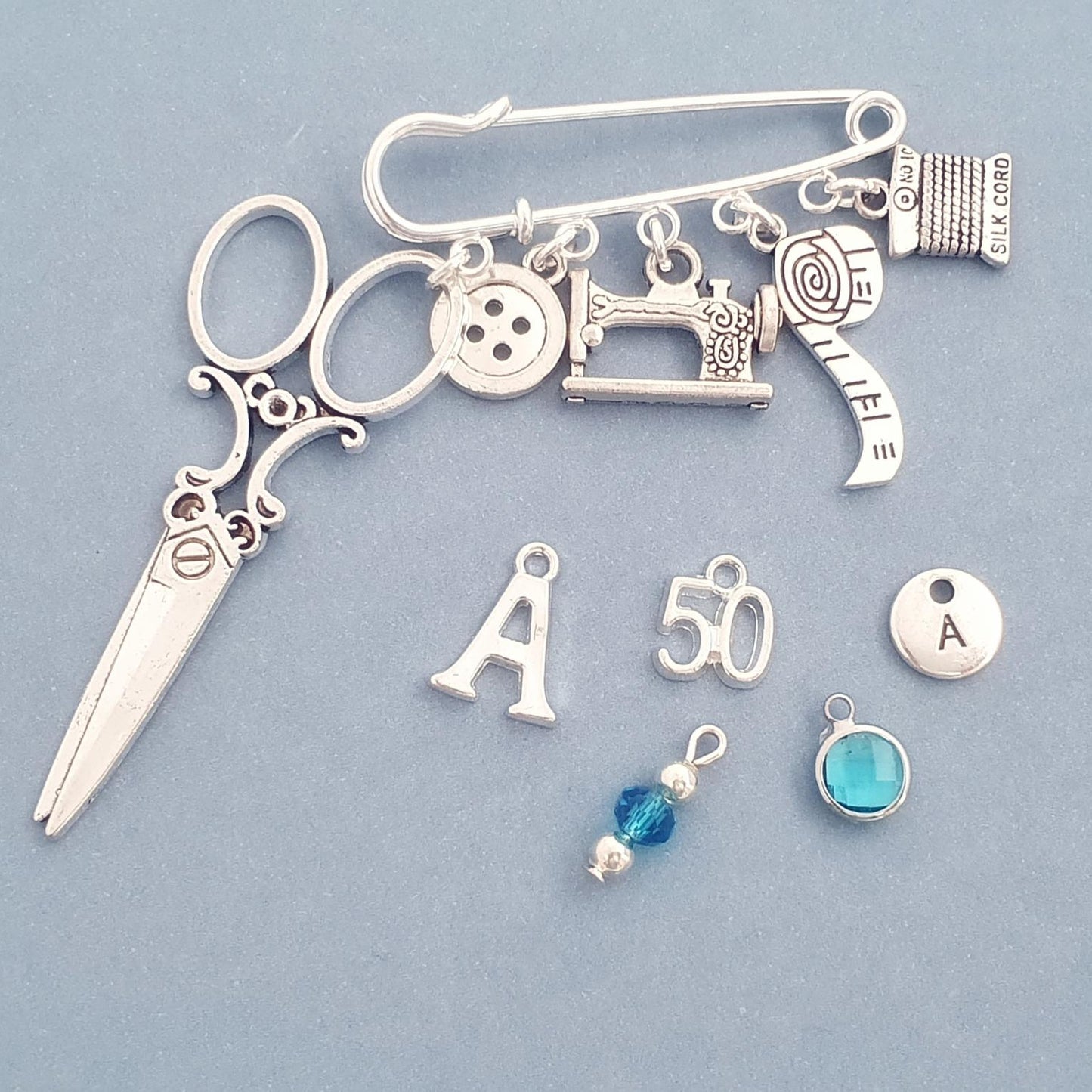Brooch with sewing themed silver charms including scissors, sewing machine, buttons, and letters on a light blue background