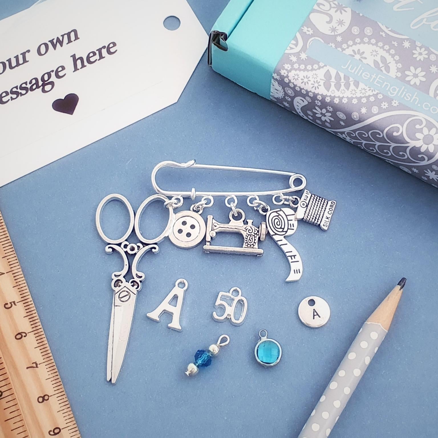 Brooch with sewing themed silver charms including scissors, sewing machine, buttons, on a blue surface with a personalized message card.
