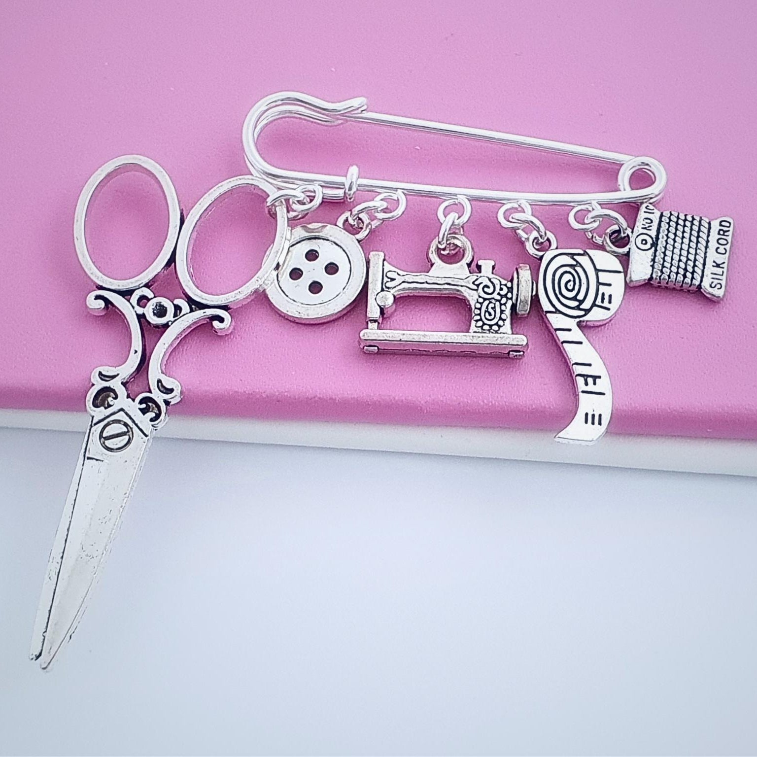 Silver brooch with sewing-themed design on a pink background