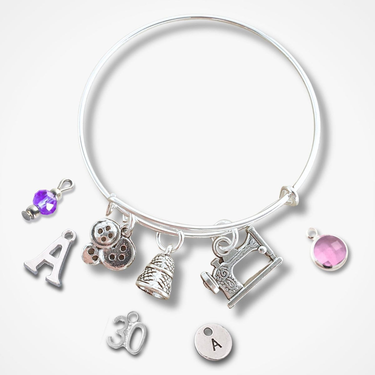 A silver charm bangle bracelet featuring various charms including a sewing machine, a thimble, a button, and letters and numbers, displayed on a clean white background.