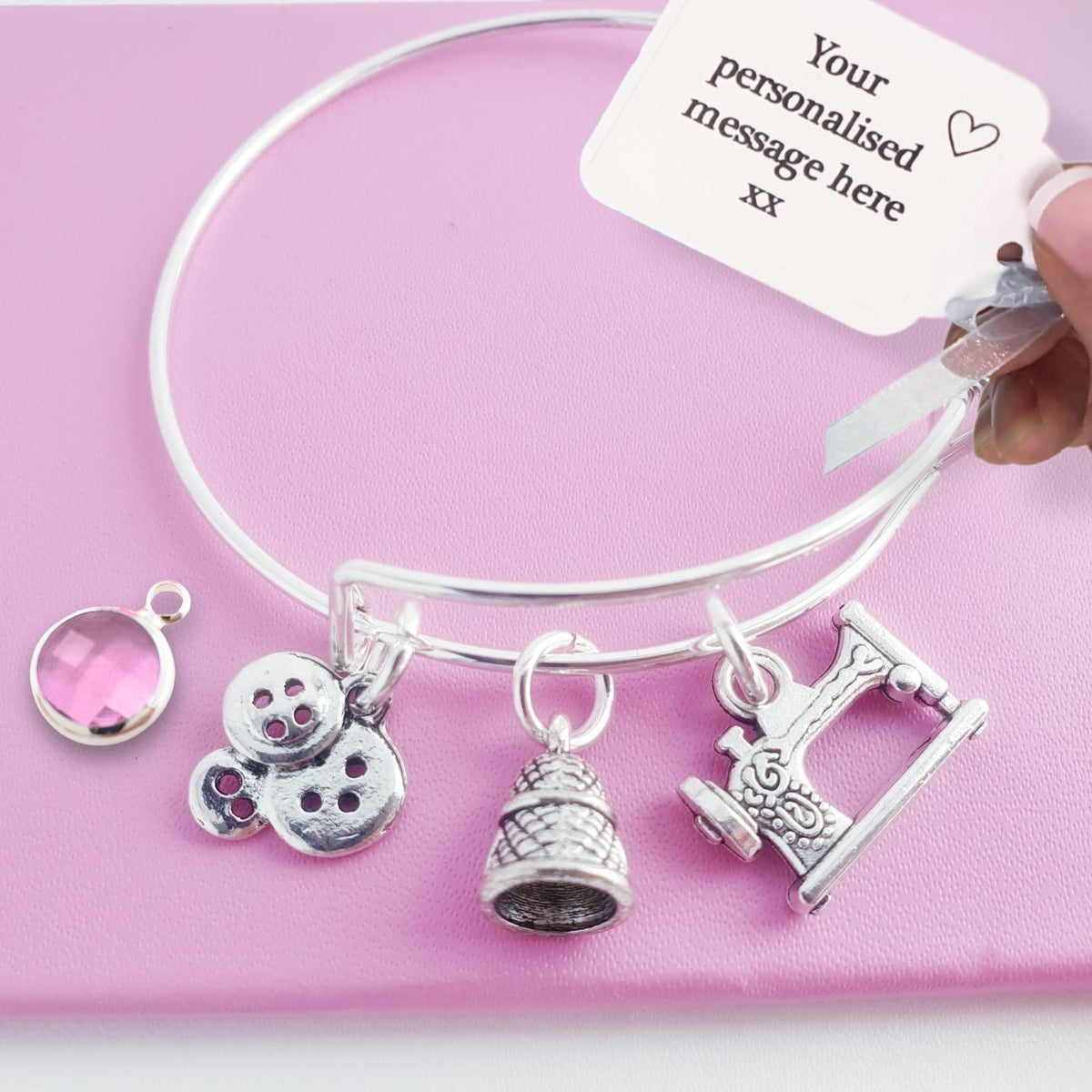 A silver bangle bracelet with four charms (a pink oval bezel, buttons, a thimble, and a sewing machine) displayed on a pink background, with a hand holding a white personalization tag.