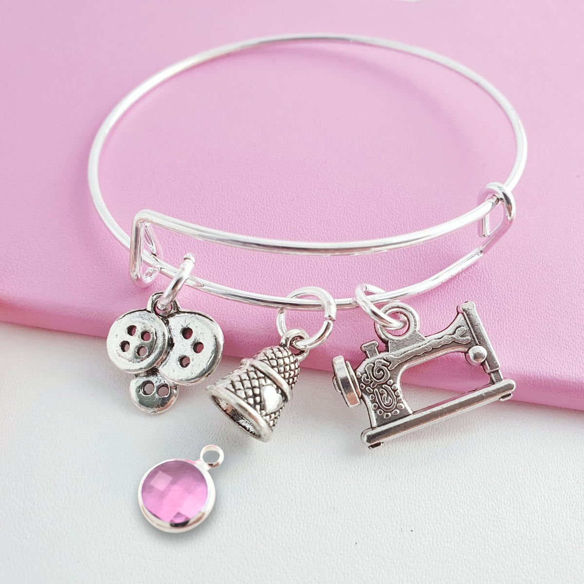 A silver-tone bangle bracelet with sewing-themed charms, including a sewing machine, thimble, buttons, displayed on a pink surface with a pink gemstone charm below.