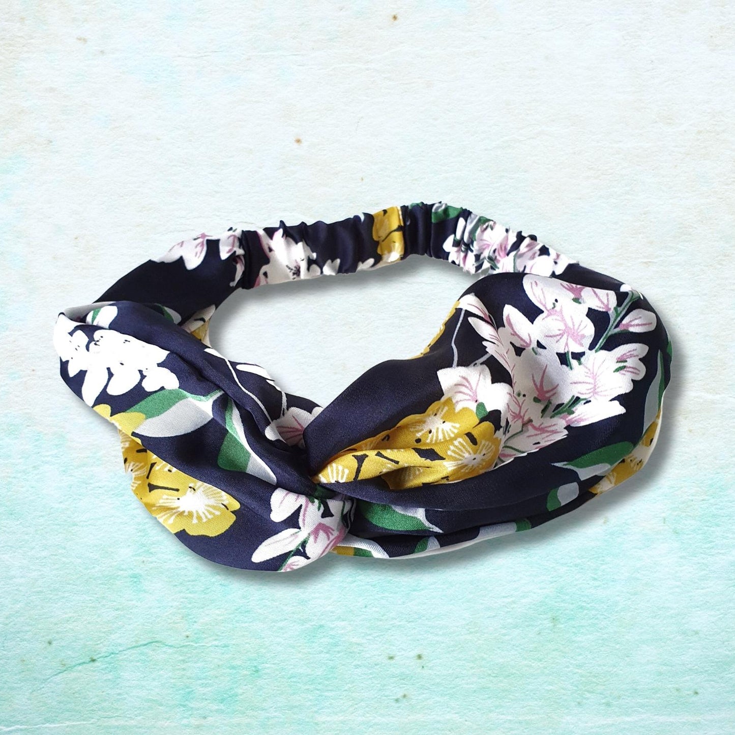 A knotted fabric headband with a floral print in navy, white, yellow, and green, displayed on a textured light blue background.