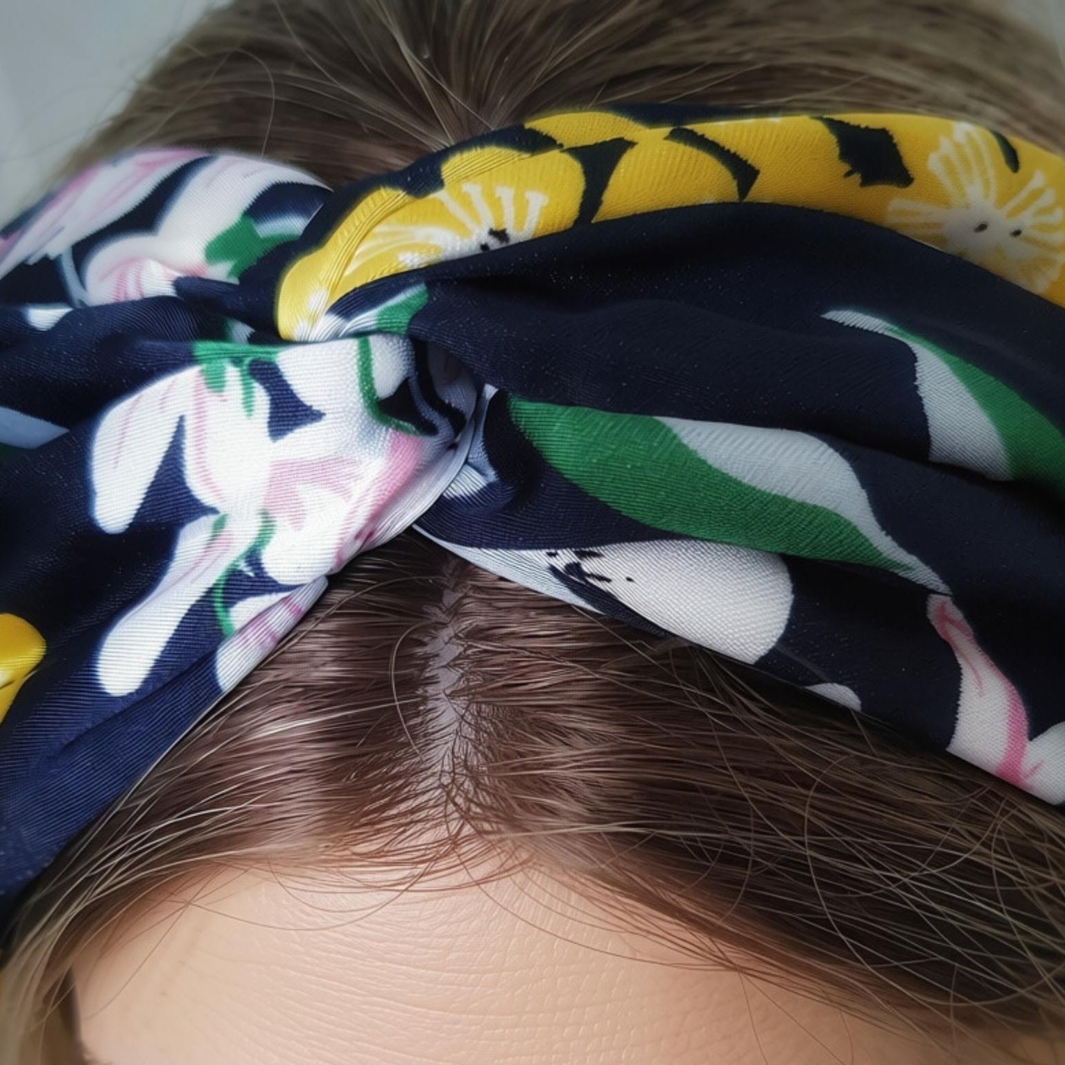 A close-up of a knotted fabric headband worn by a person, featuring a navy blue background with a floral print in yellow, white, pink, and green.