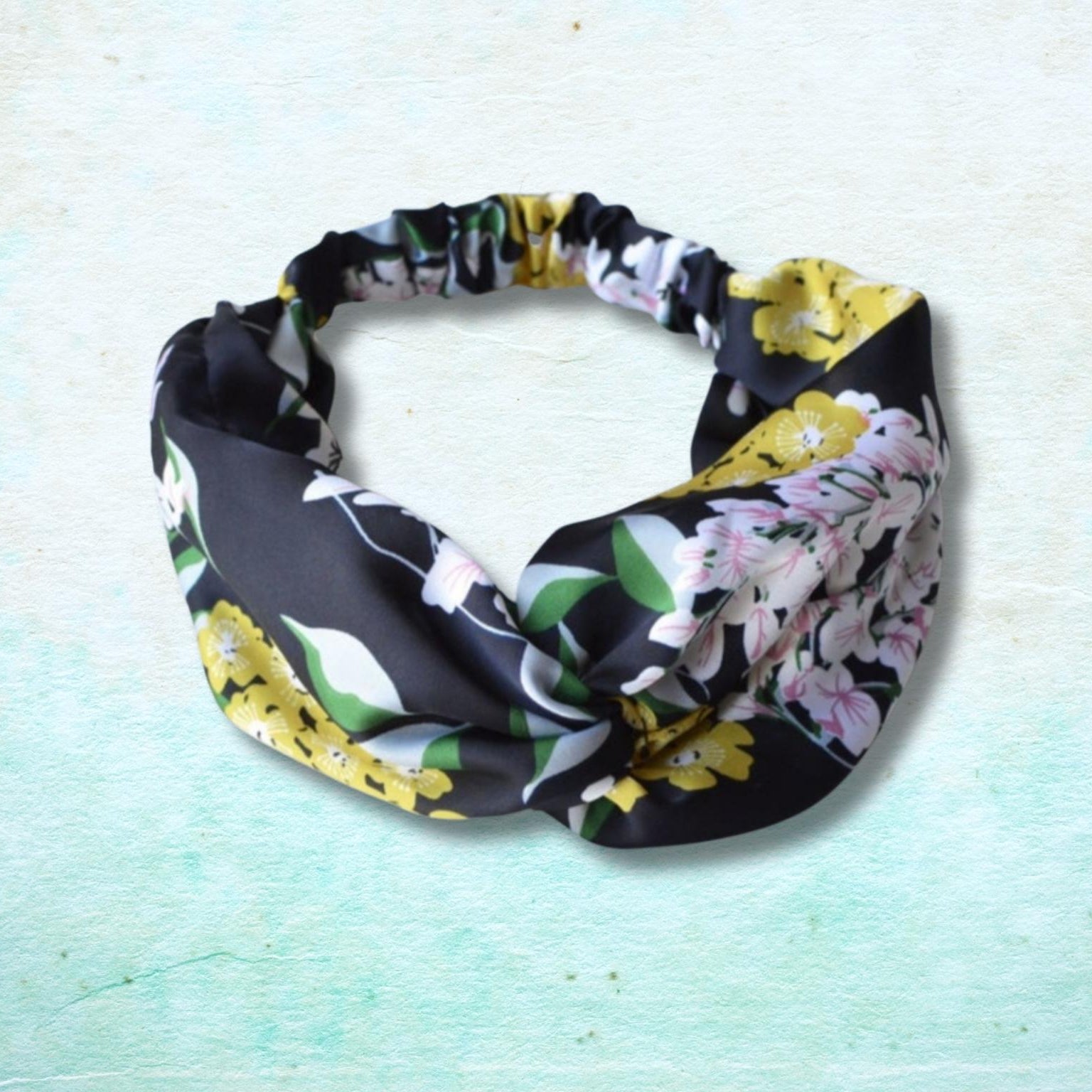 A fabric headband with a black background and a multicolor floral print featuring yellow, pink, and white flowers with green leaves, displayed on a textured light teal background.