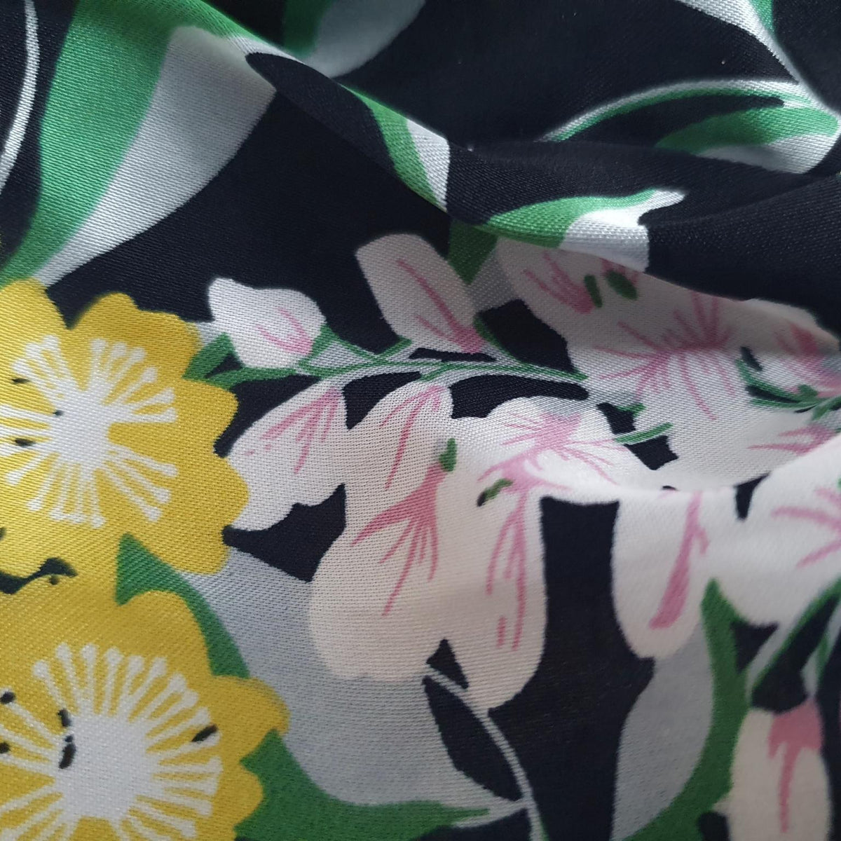 A close-up of a floral fabric with a black background, featuring yellow flowers, white flowers with pink centers, and green leaves.