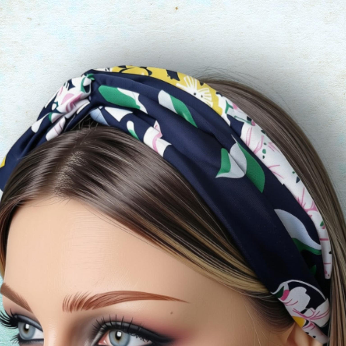 A close-up of a woman's face wearing a navy blue twisted headband with a white, green, and yellow floral pattern, set against a light blue textured background.