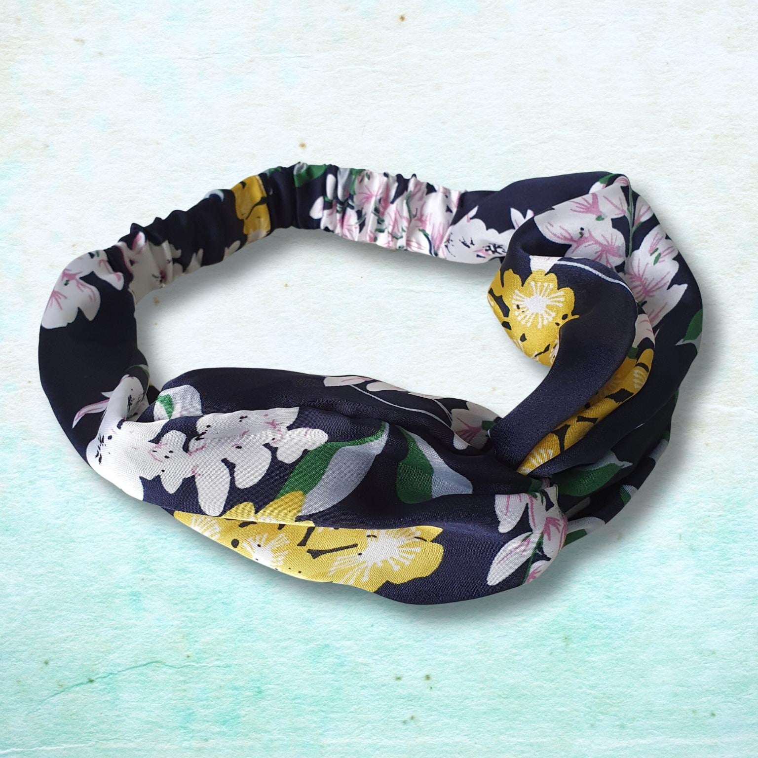 A fabric headband with a twisted knot design, featuring a blackbackground with a floral pattern of white, pink, and yellow flowers with green leaves, displayed on a textured light blue background.