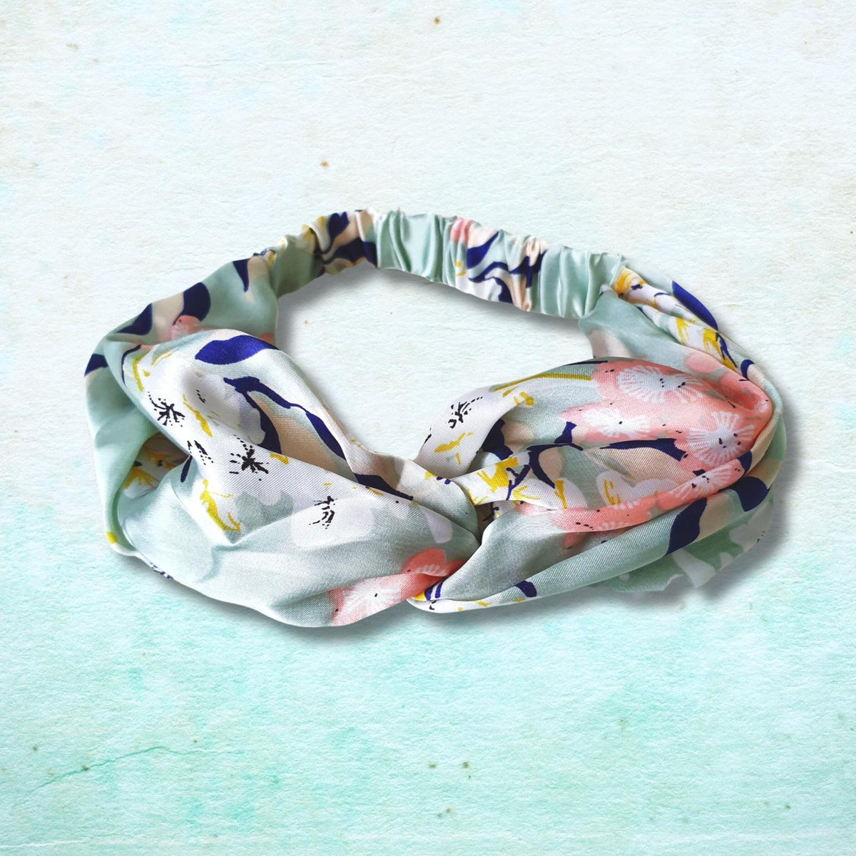 A knotted fabric headband with a multicolor abstract floral print, featuring shades of pink, blue, yellow, and green on a white base, displayed on a textured light blue background.