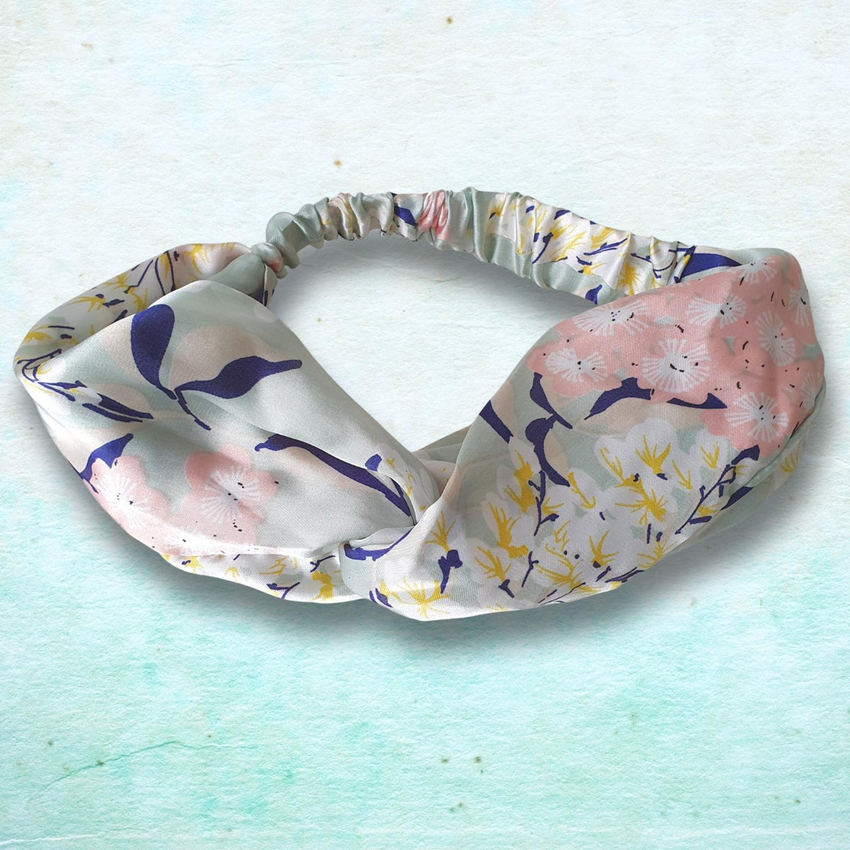 A knotted fabric headband with a floral and bird pattern in pink, purple, yellow, and white, displayed on a textured light blue background.