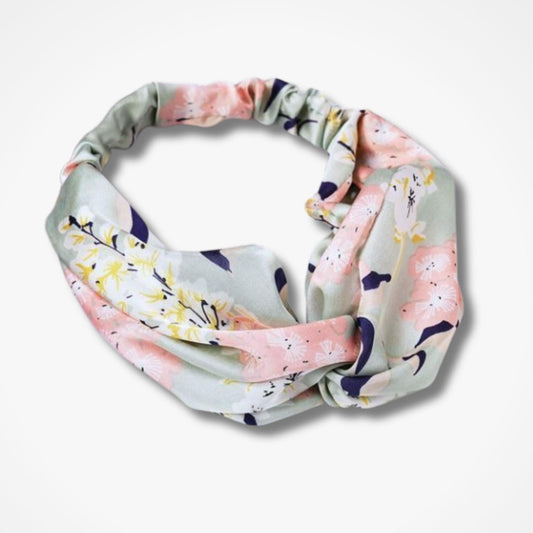 A knotted fabric headband with a pastel floral print, featuring pink, yellow, and navy blue flowers on a light sage green background, shown on a clean white studio background.