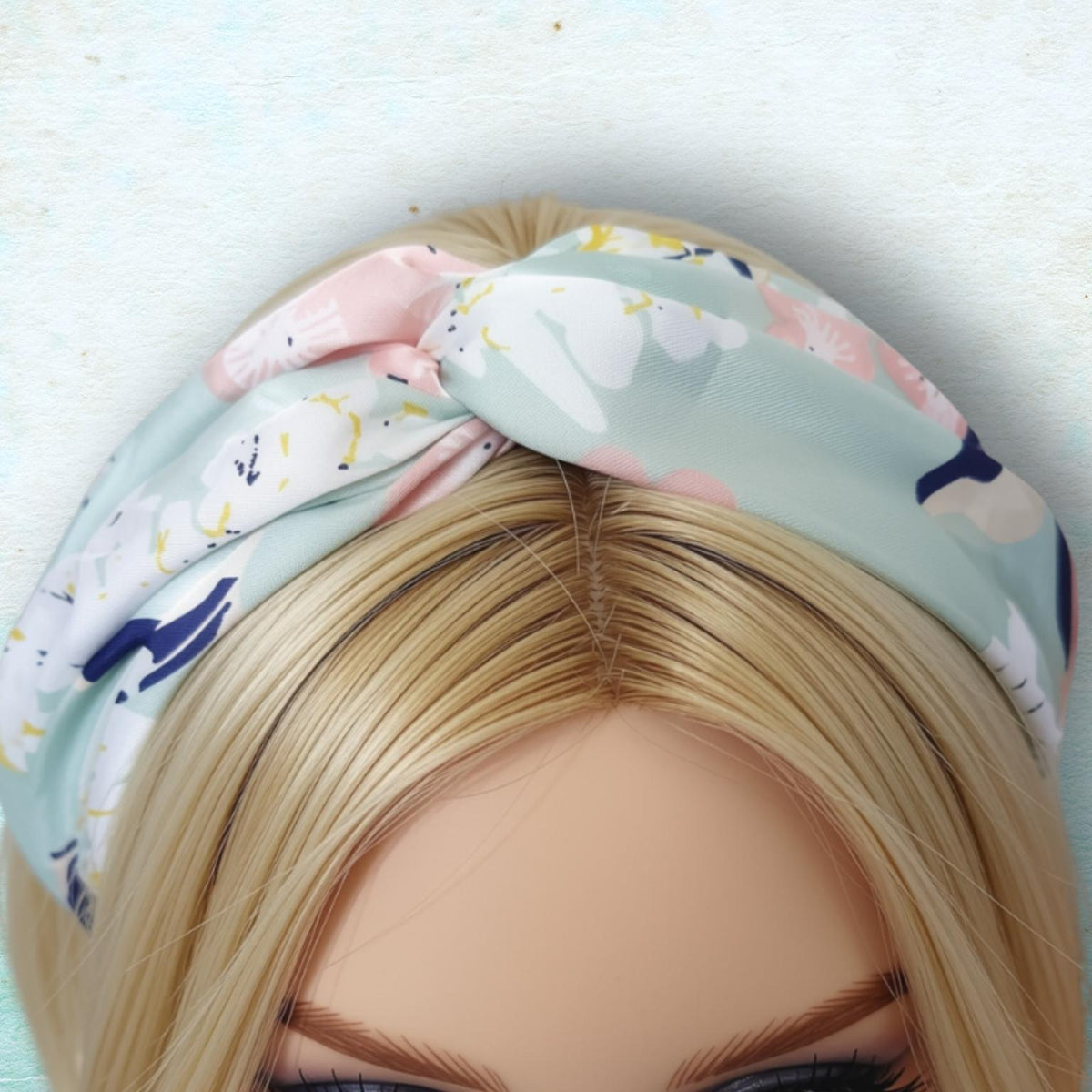 A close-up of a blonde mannequin head wearing a soft pastel headband with a pink and white, mint, and navy floral print, set against a textured light blue background.