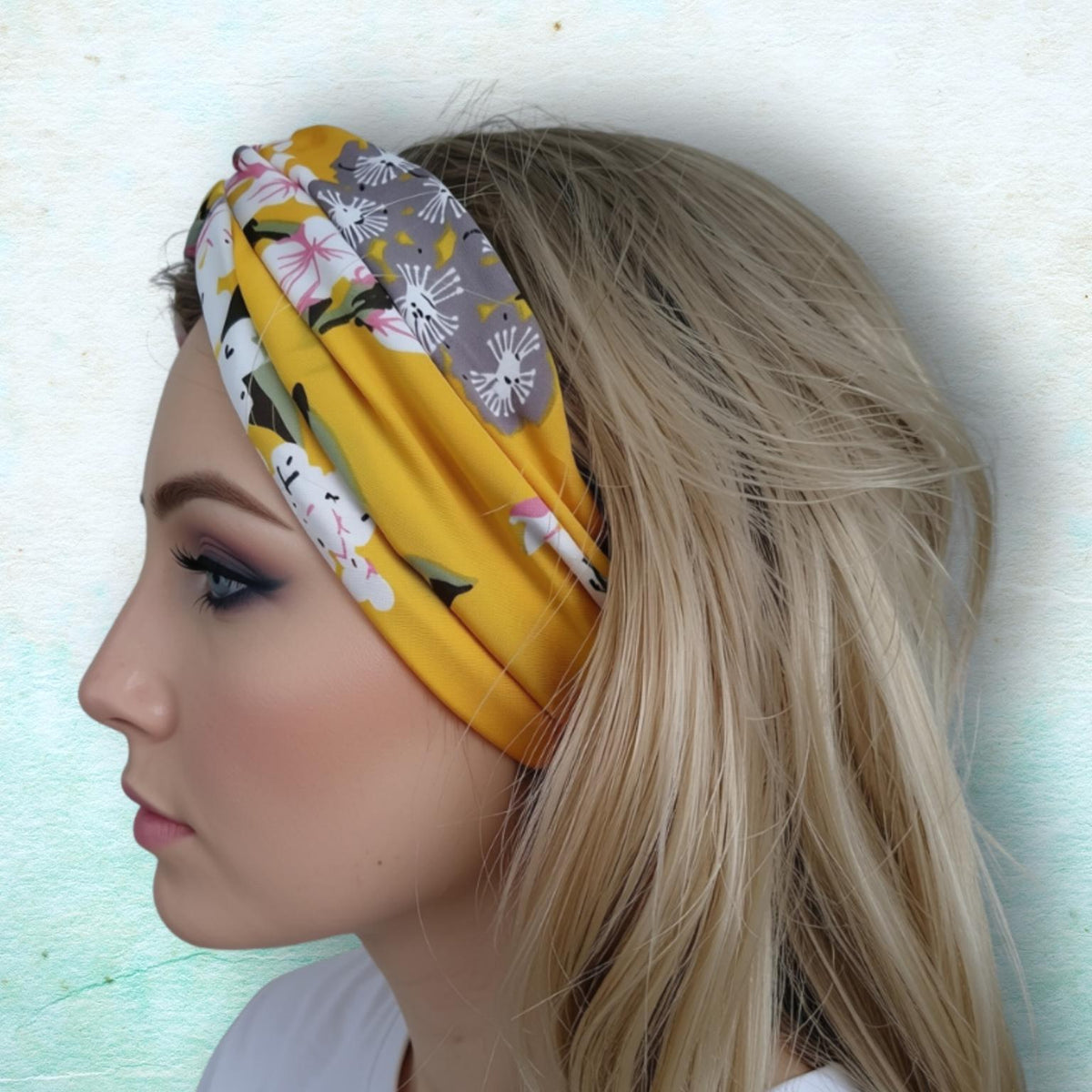 A blonde woman shown in profile wearing a mustard yellow fabric headband with a white floral print and gray patterned sections, set against a textured light blue background.