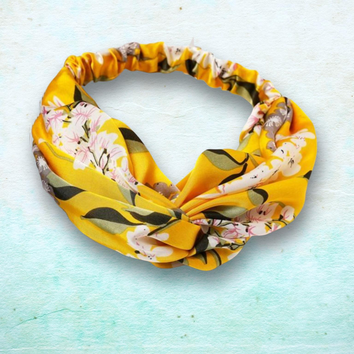 A yellow fabric headband with a floral print featuring pink, white, and green flowers, styled in a twisted knot on a light blue textured background.