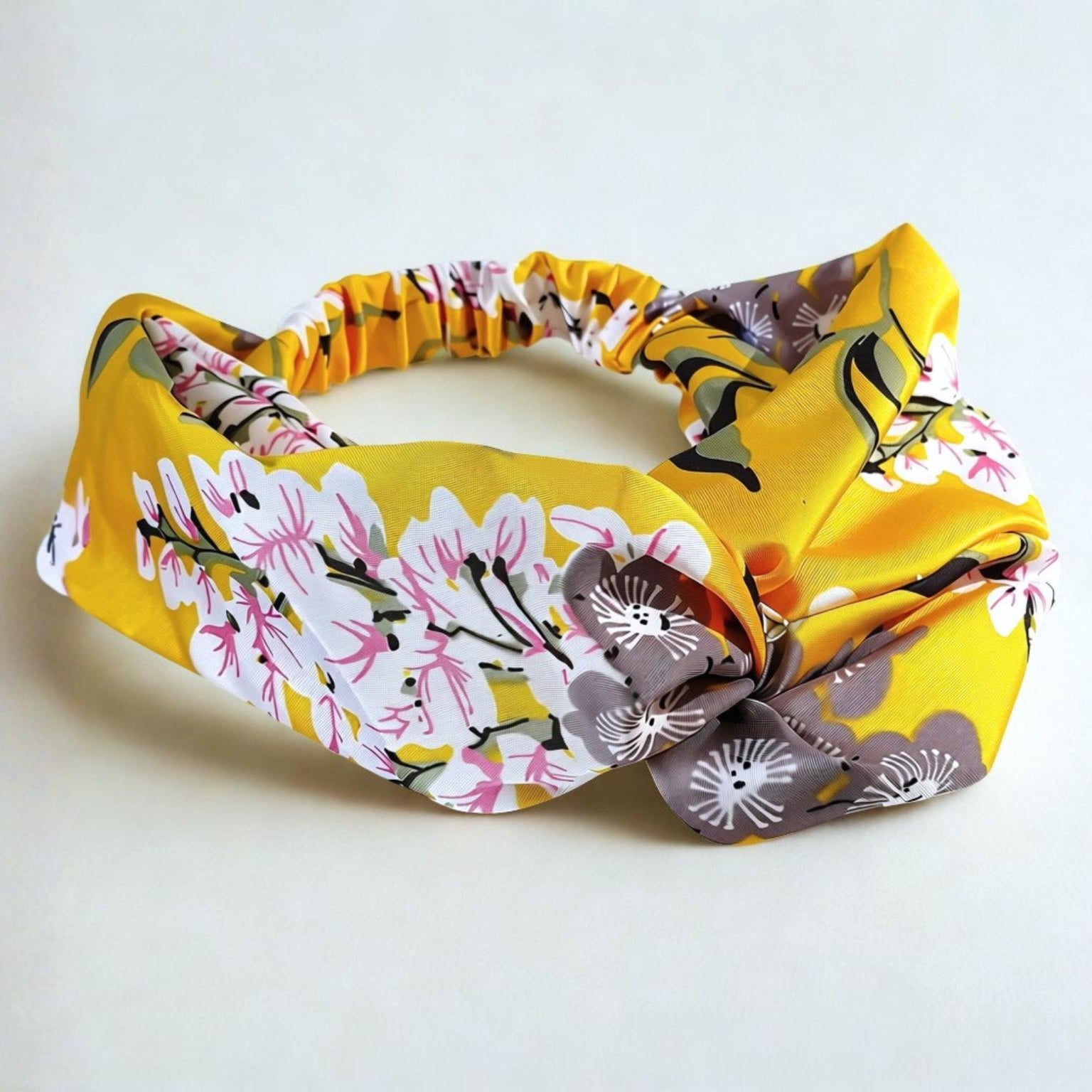 A wide fabric headband with a knotted bow, featuring a yellow background with a floral pattern of pink, white, and gray blossoms, photographed on a light off-white background.