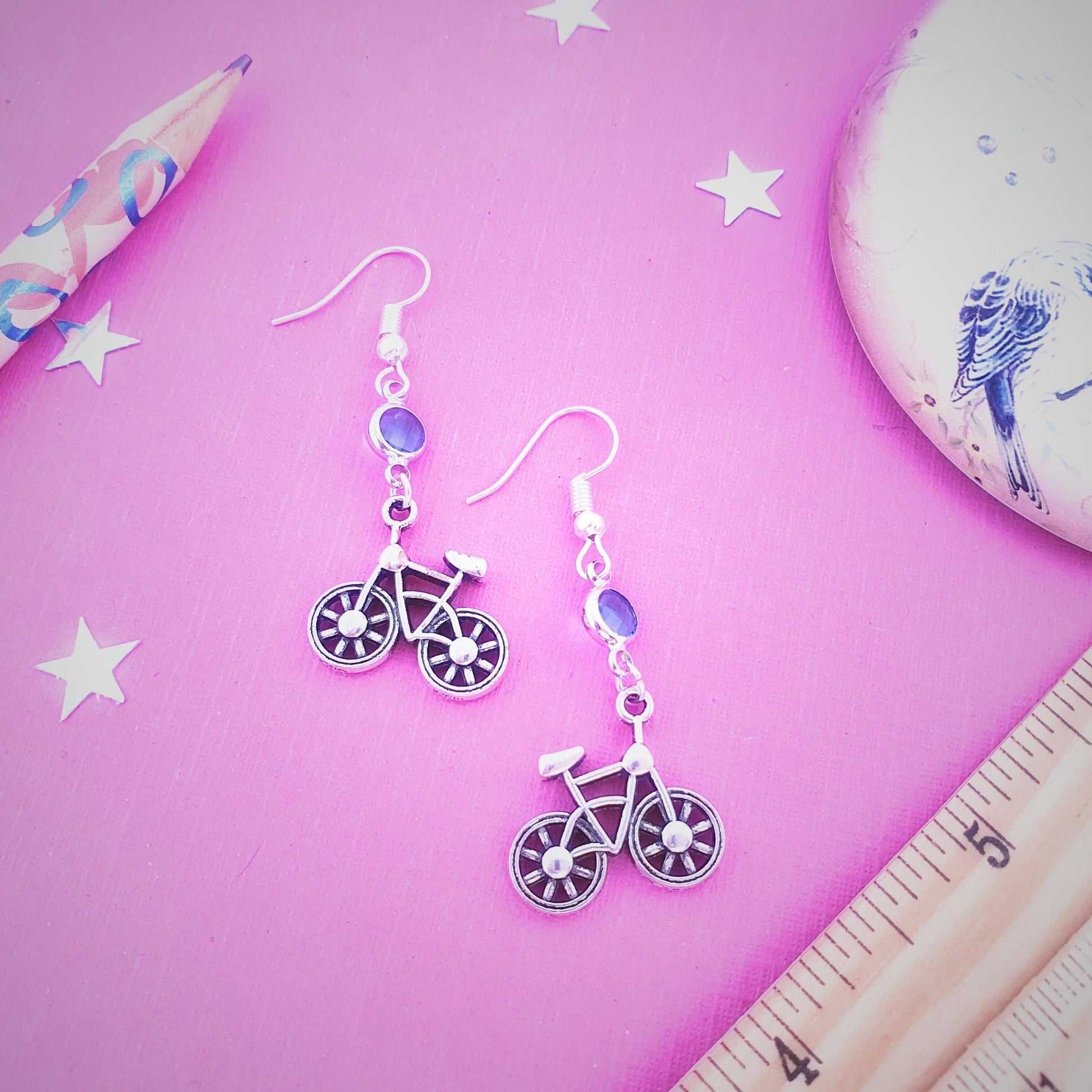 Bicycle-shaped earrings on a pink surface with a ruler and pencil.