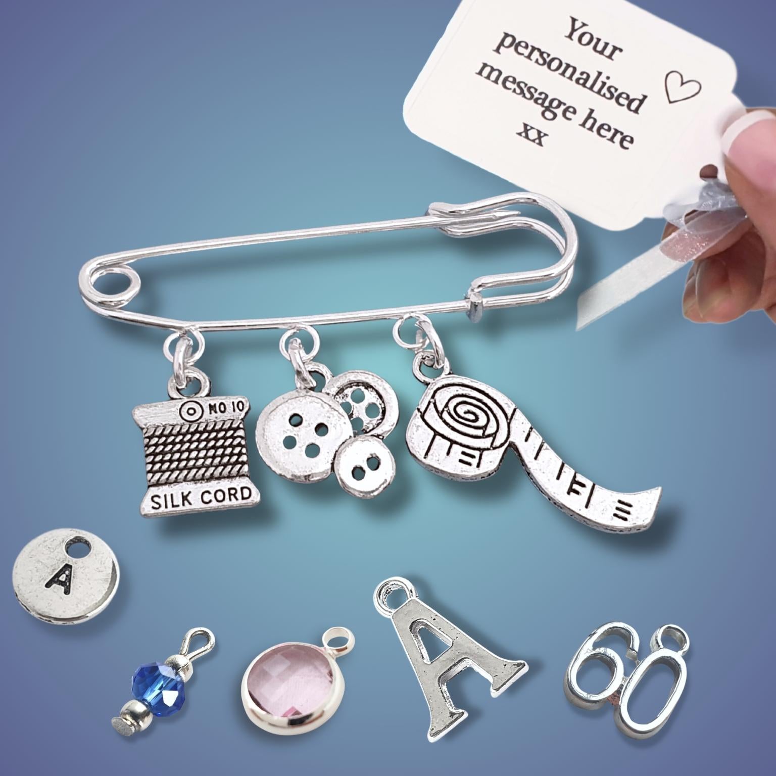 Silver charm brooch with various sewing theme charms and a hand writing on a tag against a blue background