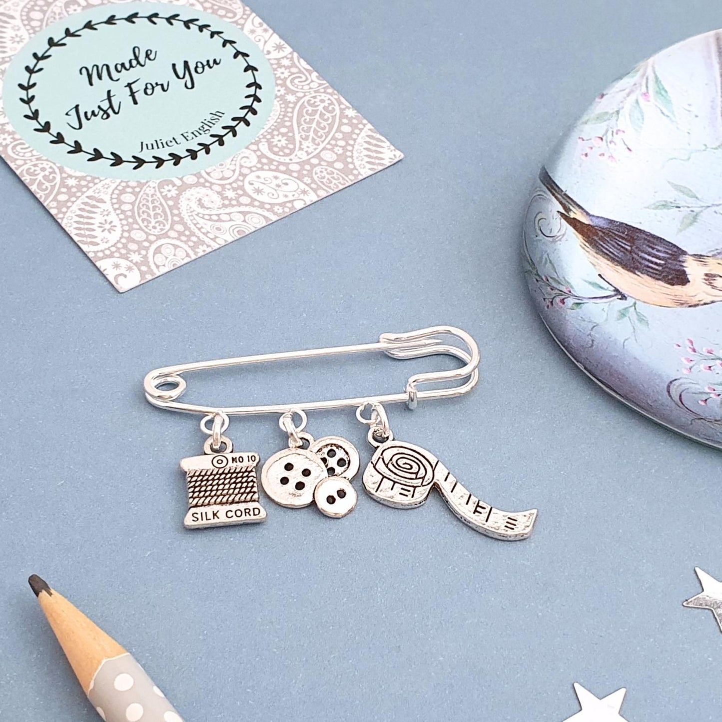 Silver charm brooch with various sewing theme charms on a blue surface with decorative elements