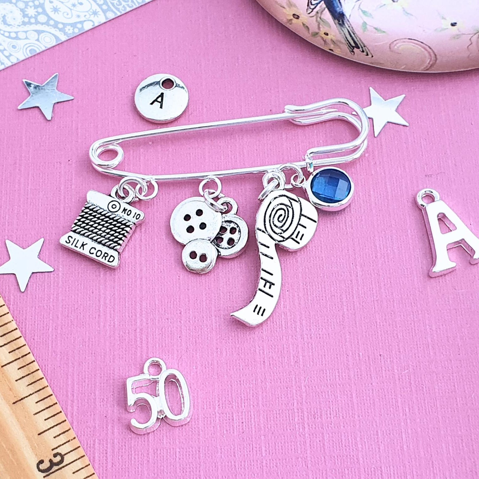 Silver charm brooch with various sewing theme charms on a pink background