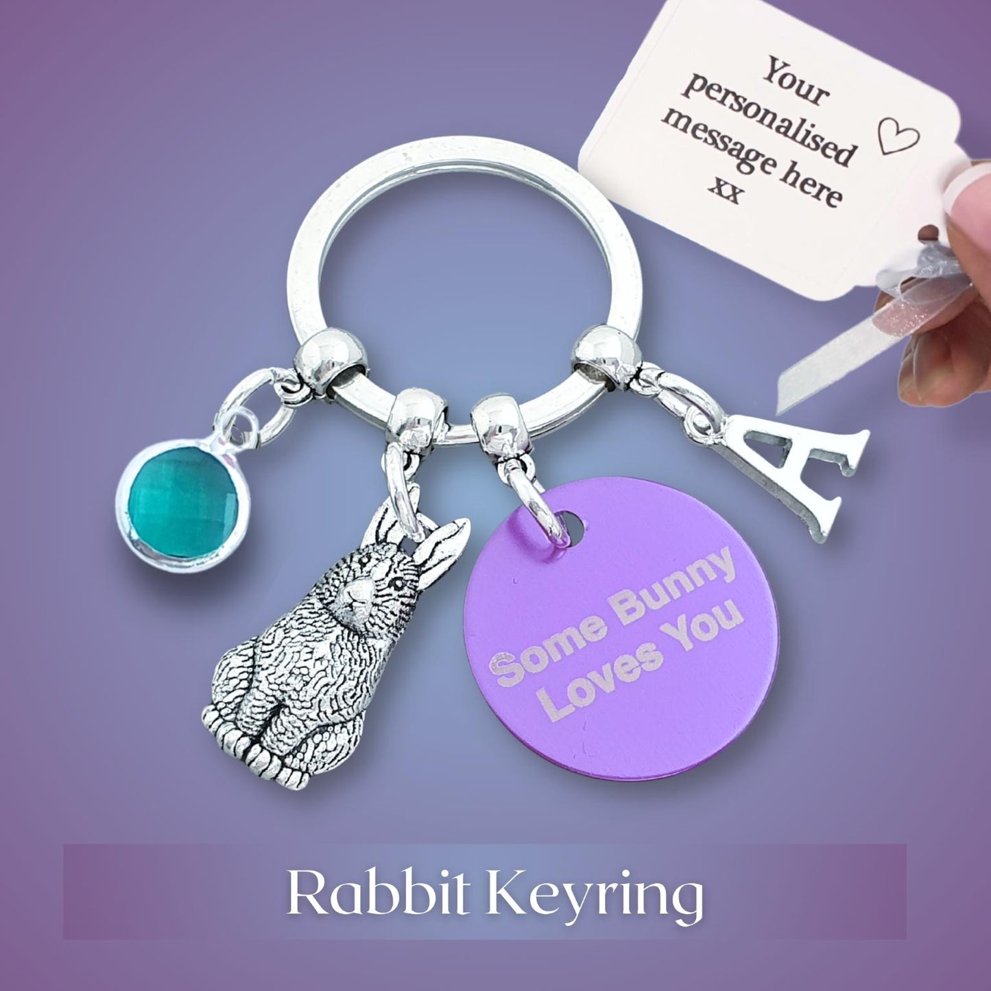 A silver rabbit keyring charm set on a purple gradient background, featuring a silver bunny charm, a round purple tag engraved with 'Some Bunny Loves You', a turquoise gemstone charm, and a silver letter 'A' charm. A hand is holding a key tag with a custom message placeholder.
