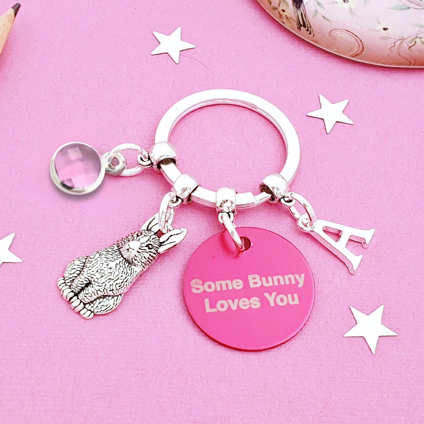 A silver keychain with a bunny charm, a pink round disc reading 'Some Bunny Loves You', and a letter 'A' charm, displayed on a pink background with decorative star cutouts.