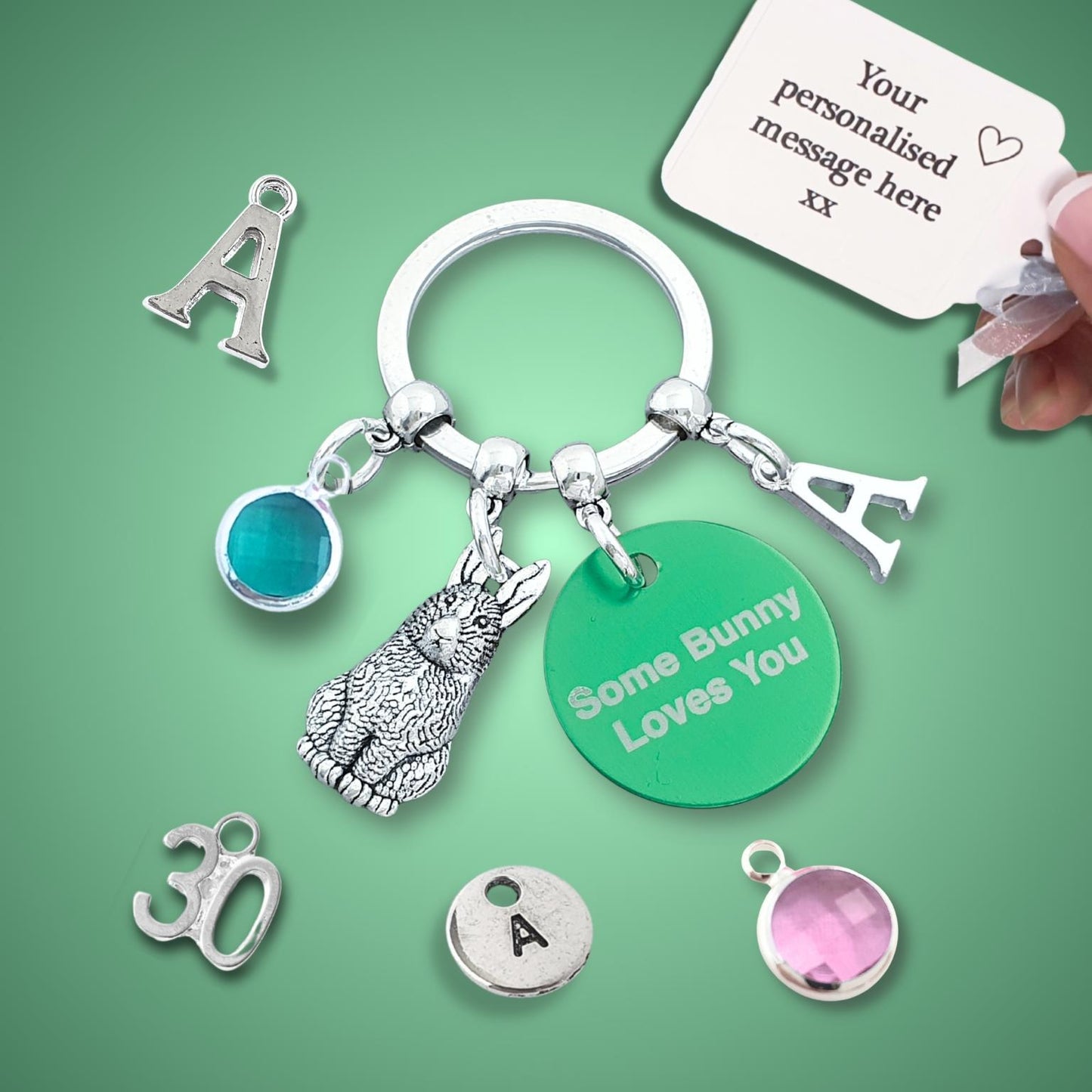 A silver keychain set with customizable charms, including a bunny charm, a green round charm reading 'Some Bunny Loves You', and letter 'A' charms, displayed on a green background with a hand holding a personalization tag.