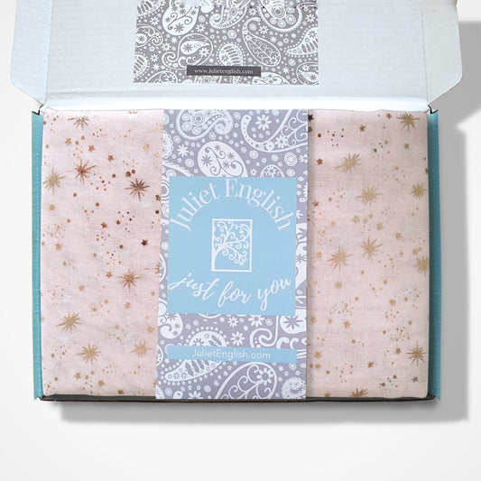 An open gift box containing a folded fabric scarf with celestial patterns in soft pink and rose gold, with a central card reading 'Juliet English just for you'.