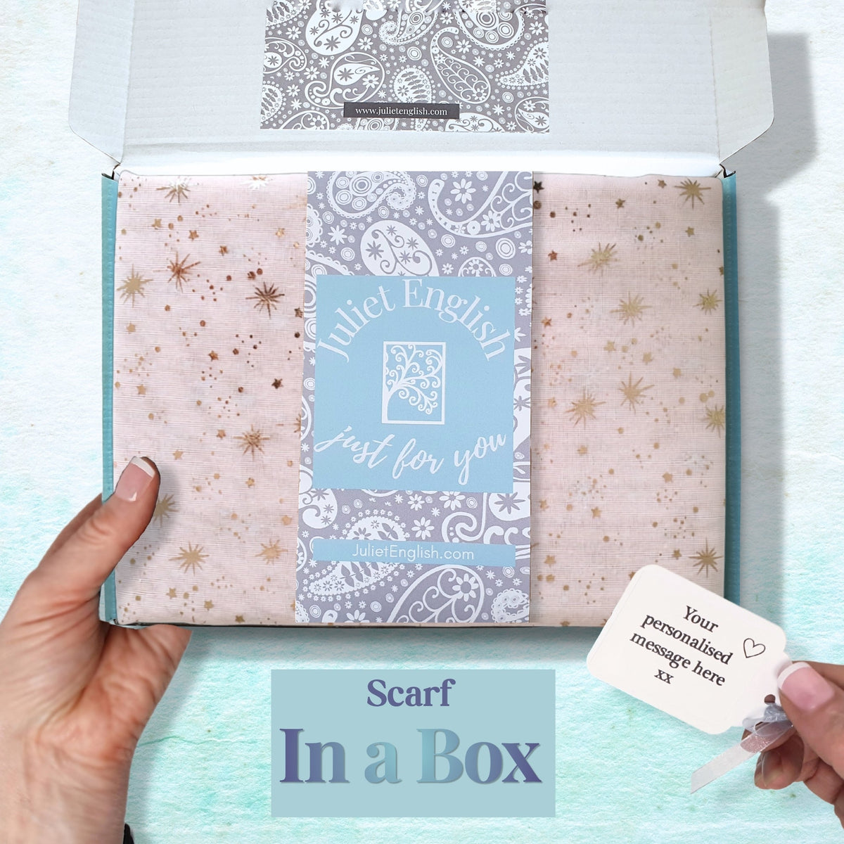 A hand holds an open box containing a folded scarf in pale pink  with rose gold star prints. A central blue label reads 'Juliet English just for you'.